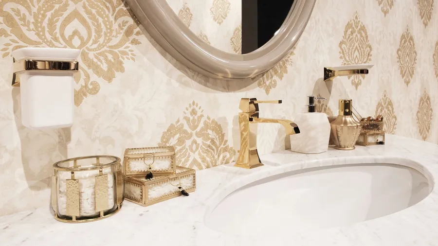 Bathroom interior featuring a gold faucet, soap dispenser, decorative glass boxes with gold accents, and a candle holder on a marble countertop.