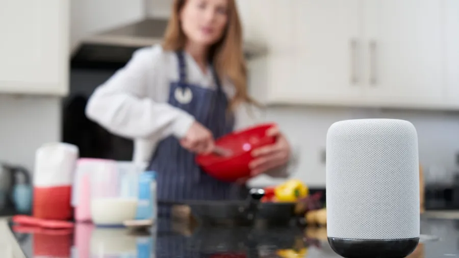 A woman in a kitchen, holding a red bowl and preparing food, with a smart speaker in the foreground.