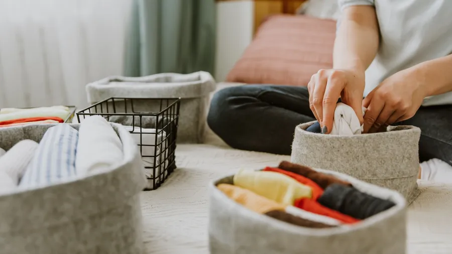 A person sorting folded clothes into gray baskets on a bed.