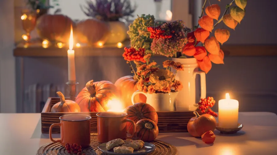 Two orange cups of tea and autumn decor with pumpkins, flowers and burning candles on table.