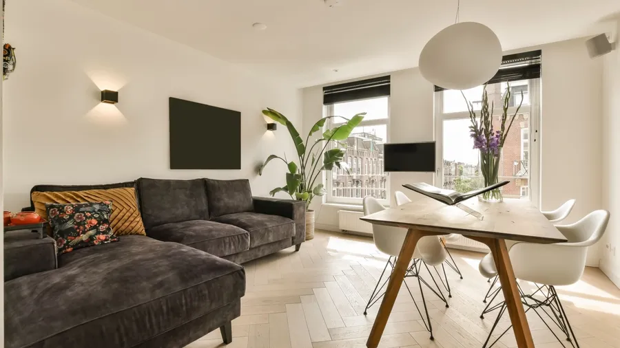 A contemporary white living room interior with a dark sofa, wooden table, and large windows.