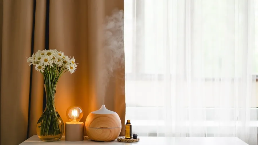 An aromatic oil diffuser, essential oil, flower vase, and an Edison bulb lamp on a white table near a window with sheer curtains.