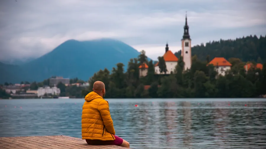 Traveler man sitting on the lake wooden pier against island with church of Lake Bled in Slovenia. Travel lifestyle concept.