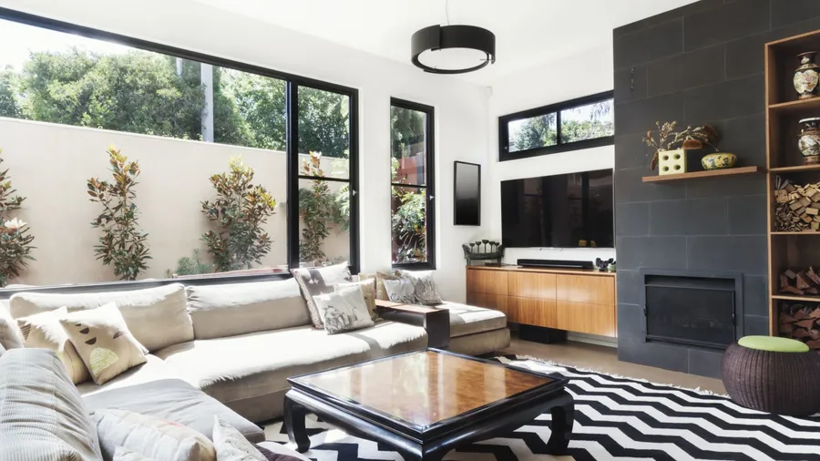 A modern living room with large windows, a beige sofa, and a black and white patterned rug.