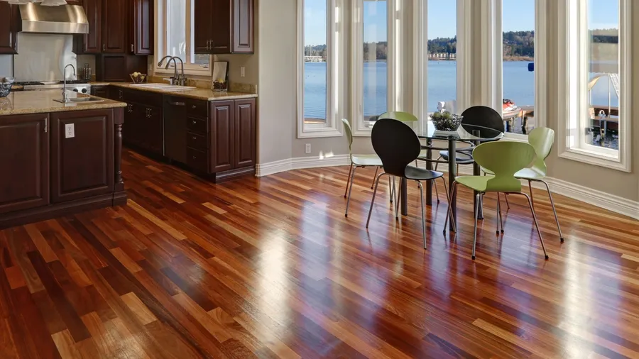 A modern kitchen with rich wooden cabinets, large windows overlooking the water, and a dining table set with colorful chairs.