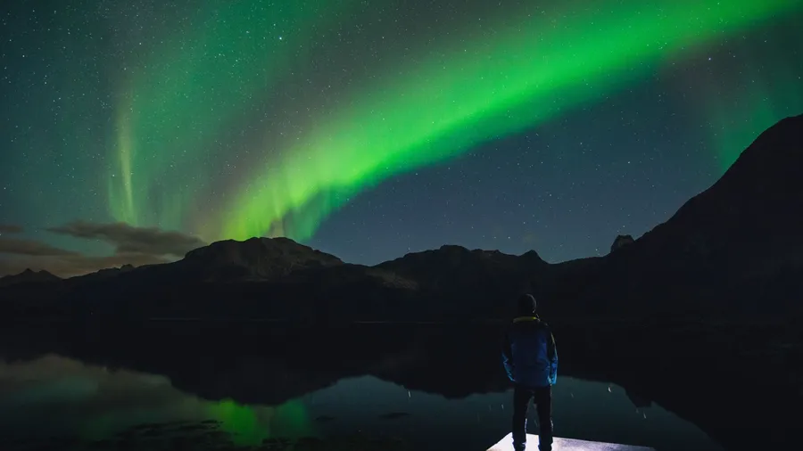 Person stands on dock beneath vivid green Northern Lights, mirrored in calm water under star-filled sky.