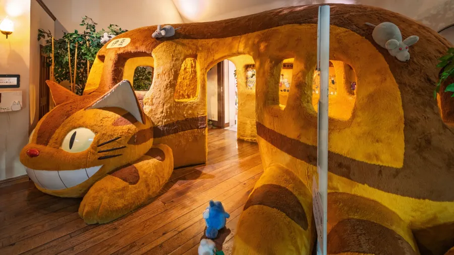 A colorful, plush cat bus structure from Studio Ghibli's "My Neighbor Totoro," with small stuffed animals sitting atop it on a wooden floor.