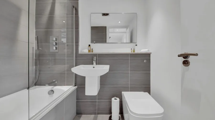 A stylish bathroom interior with gray tiled wall, glass-enclosed bathtub, sleek white sink, floating toilet, and a mirror on a white wall.