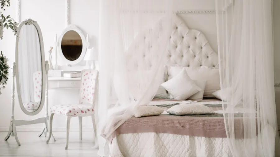 A bedroom interior with a bed with throw pillows and a canopy, a dressing table and chair, and a full length mirror near the bed.