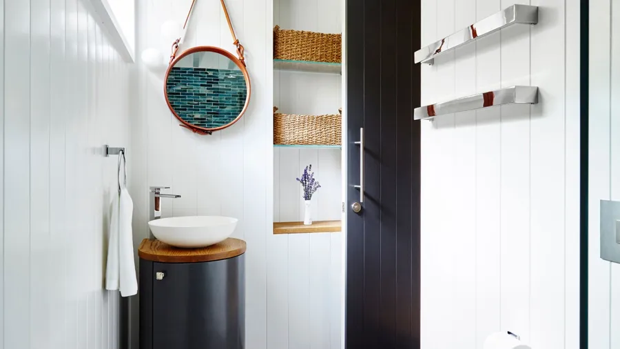 A small bathroom interior with round mirror, curved vanity, and woven baskets on glass shelves.