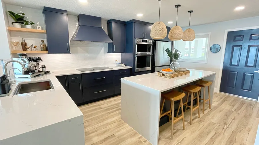 A modern kitchen interior with navy blue cabinets, recessed lighting, pendant lights, and marble countertop.