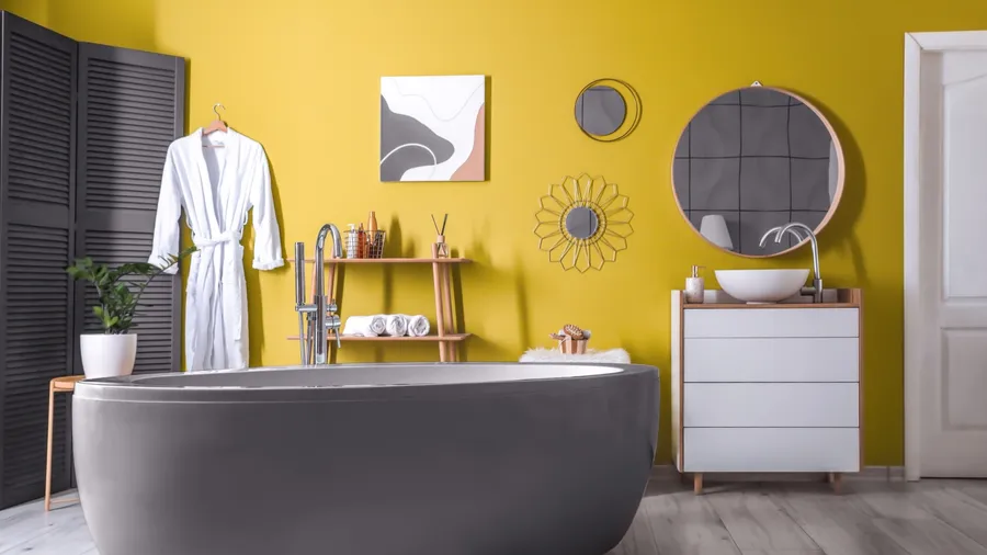 A modern bathroom with a yellow wall, a gray bathtub, a potted plant, a wooden table, shelves, and wall art.