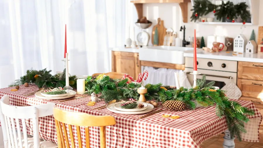 A dining table setting with checkered tablecloth, pine leaves garland centerpiece with pinecones and candycanes, candle holders with white and red candles, and dinnerware.