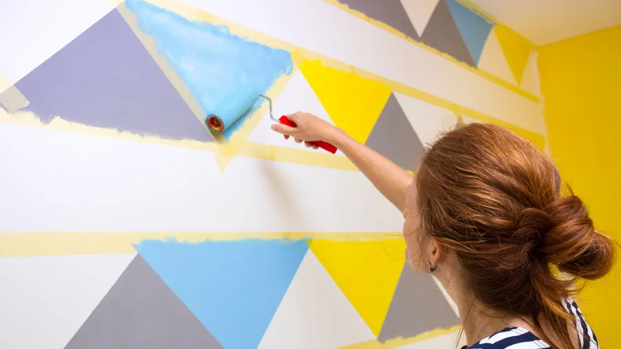 A woman painting geometric patterns on the wall using masking tape and roller.