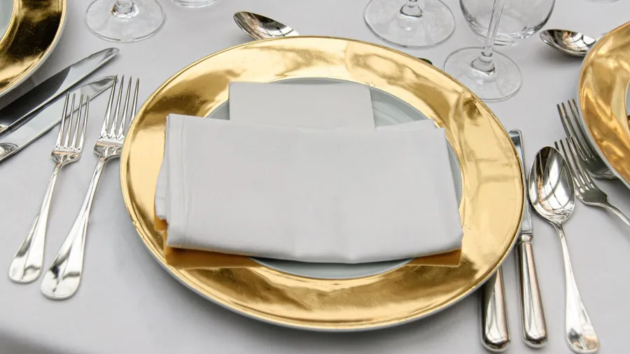 A formal table setting with a gold-rimmed plate, white napkin, and silver cutlery.