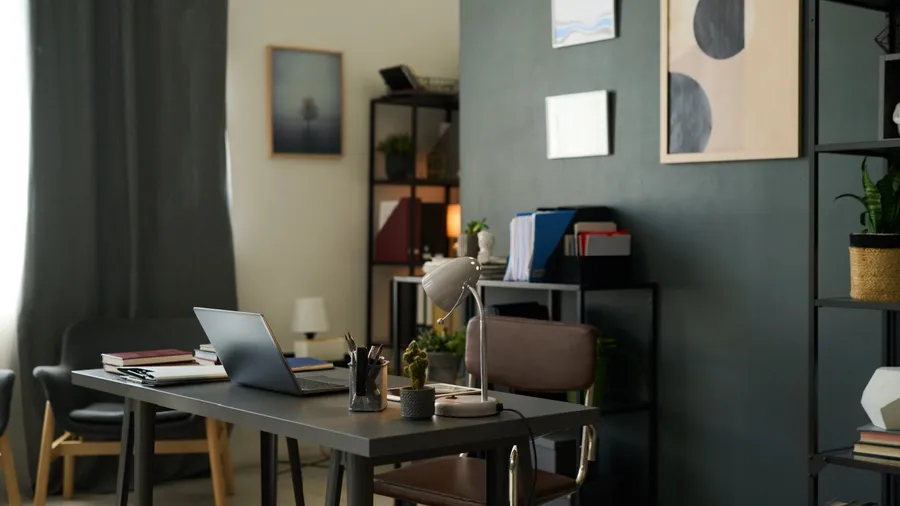 A modern home office with a desk, laptop, lamp, books, and shelves with various items.