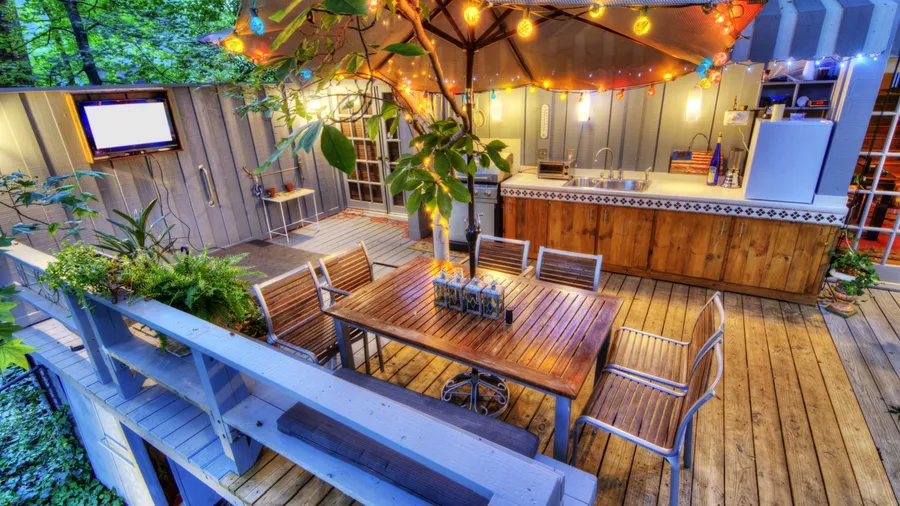 An outdoor deck with wooden furniture, a large umbrella, string lights, and potted plants near an outdoor kitchen.