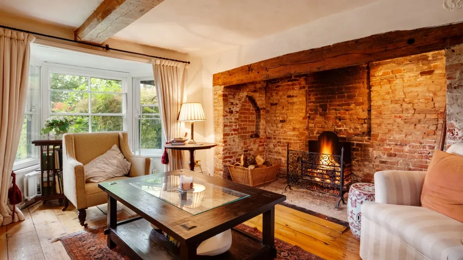 A cozy countryside living room with a rustic brick fireplace, wooden beams, soft lighting, and vintage furniture near a large bay window with cream curtains. A cozy countryside living room with a rustic brick fireplace, wooden beams, soft lighting, and vintage furniture near a large bay window with cream curtains.