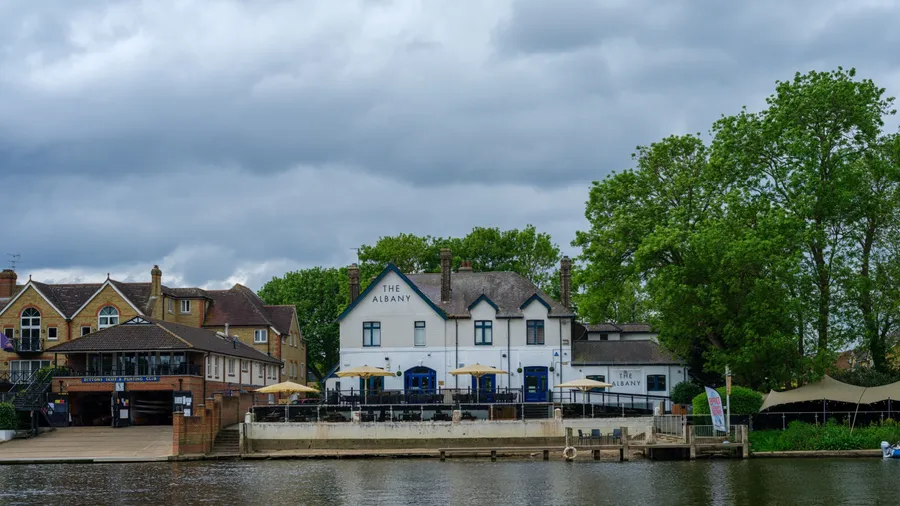 UK, Hampton Court, 05 May 2025 Riverside buildings with outdoor terraces face the calm Thames waters near Hampton Court, offering scenic views and relaxed dining by the river.