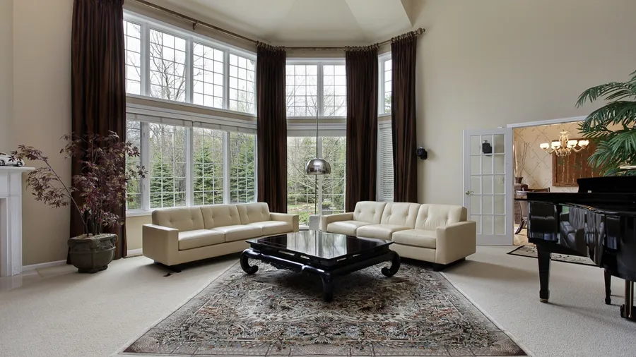 A spacious living room with beige sofas, a black coffee table, a patterned rug, large windows, and a grand piano.