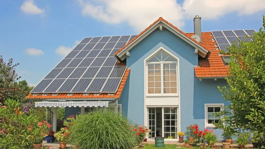 Blue house with red roof tiles, solar panels, and a lush garden filled with colorful plants and flowers.