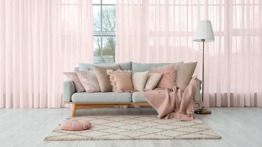 A living room interior with a cozy sofa with throw blanket and pillows, floor lamp, blush pink sheer curtains on the windows, and a rug.