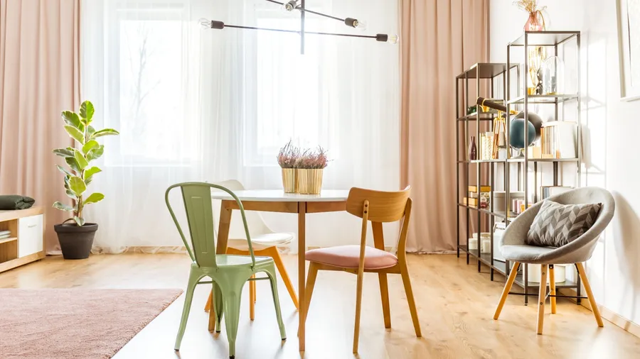 A modern living room with a round wooden table, three different chairs, a potted plant, a bookshelf, and a cushioned chair.