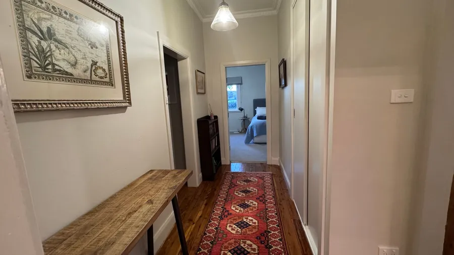 A hallway with a wooden floor, a red runner rug, a wooden console table, framed artwork on the walls, and a view into a bedroom.