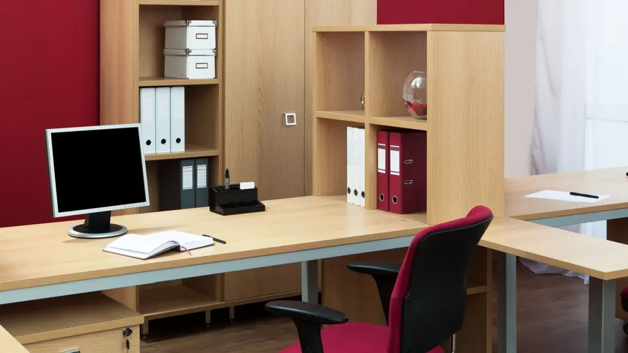 A modern office workspace with wooden desks, a computer monitor, office supplies, and shelves containing binders and storage boxes.