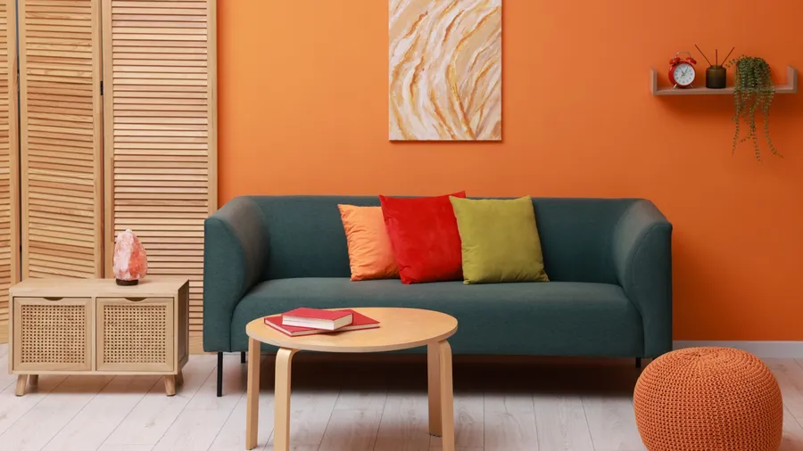 A living room interior with a cozy sofa, throw pillows, wooden coffee table and side table, and floating shelf and artwork on the orange wall.