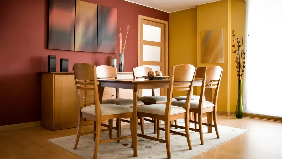 A dining room interior with table, chairs, rug, green vase, and artwork on the wall.