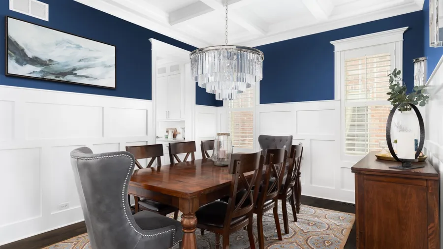 A dining room with wooden table, chairs, rug, a crystal chandelier hanging from the ceiling, and wainscoting on a navy wall.