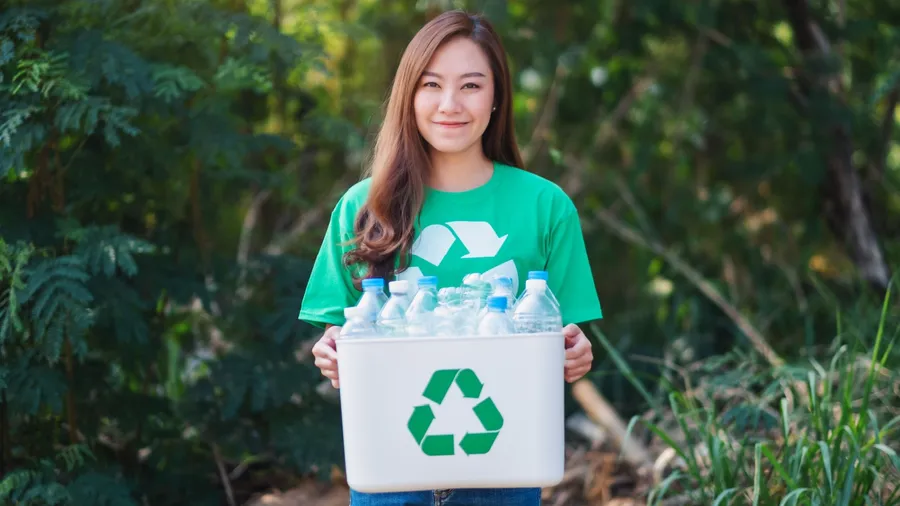 A woman holding a recycling bin filled with plastic bottles, wearing a green shirt with a recycling symbol, standing in a natural outdoor setting.