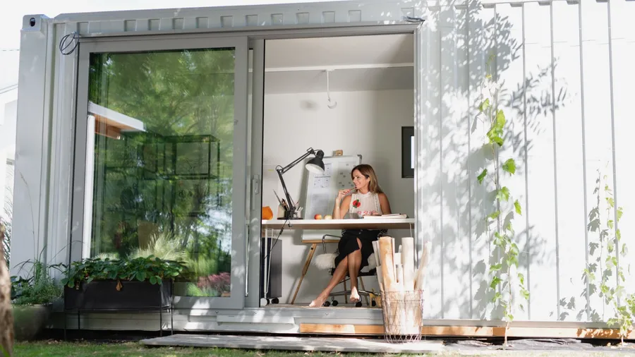 A modern container office surrounded by greenery with a woman working at a desk inside.