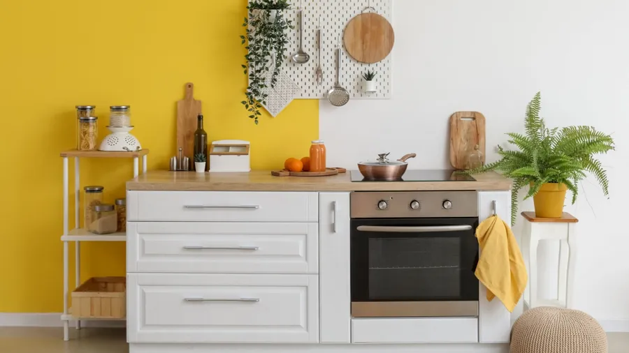 A kitchen interior with white drawers, oven, plants, a pegboard holding utensils, cutting board, and a spice rack on a mustard yellow wall.