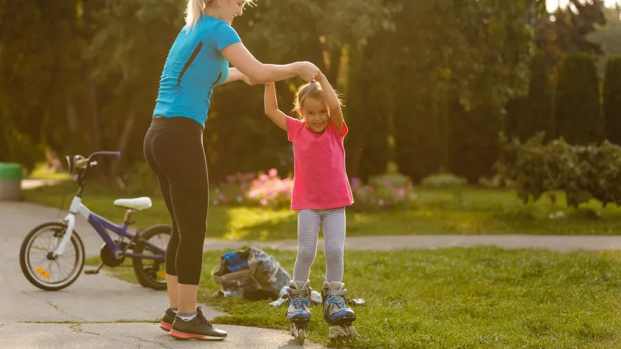 A young girl in a blue shirt helping a smiling girl in a pink shirt to skate outdoors in a park at sunset.