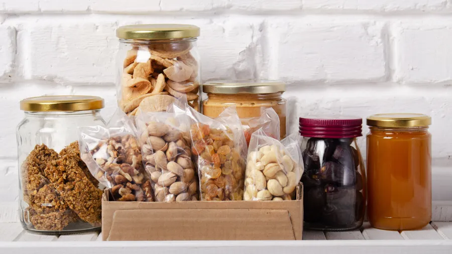 Different dry fruits and snacks in a box and glass jars placed in front of a white wall.