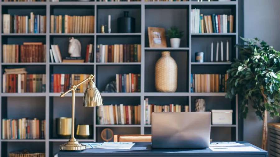 A home office interior with laptop and lamp on a desk in front a bookshelf with decor accents and books.