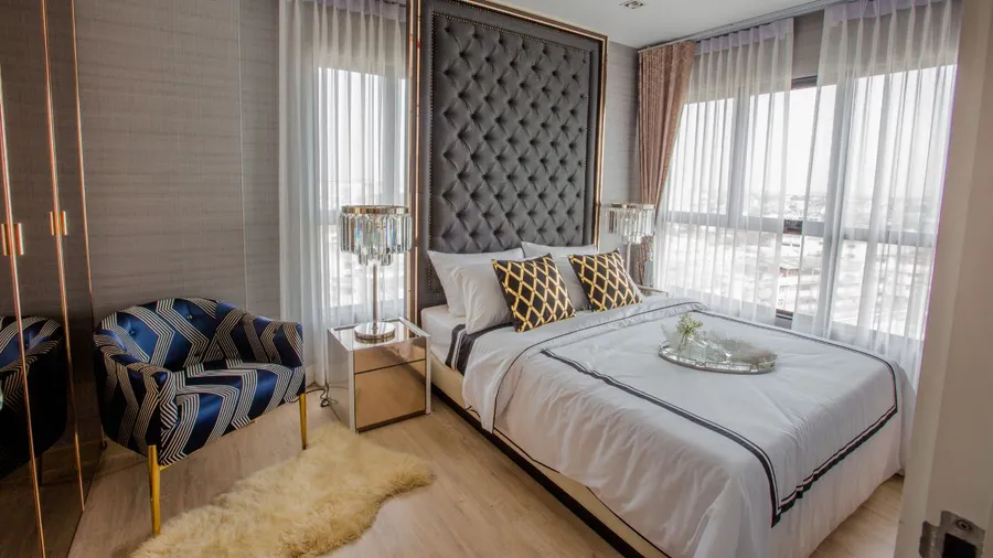 A stylish bedroom with a large bed, tufted headboard, decorative pillows, and large windows with sheer curtains.