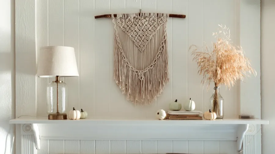 Thanksgiving home decor - white wooden fireplace with wall macrame, cream and beige pumpkins, pine cones garland and rustic dry wheet bouquet. House fall decoration.