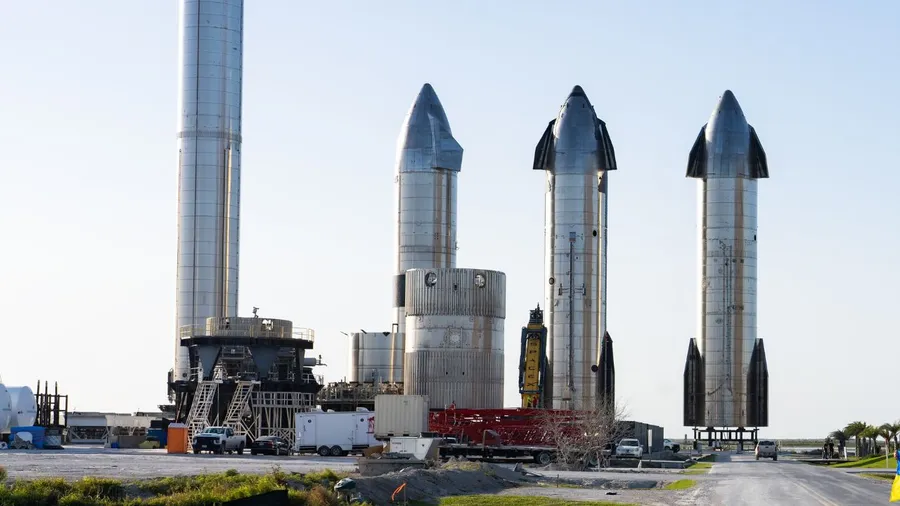 A row of SpaceX Starship rockets standing at the launch site in Boca Chica, Texas. The rockets are towering, with shiny metal exteriors, ready for testing or launch preparations.