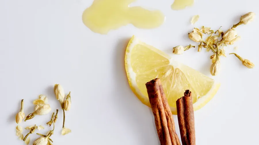 Top view of oil flowing out next to slice of lemon, sticks of cinnamon and vanilla buds on white background.