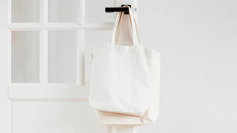A white tote bag hanging on a door with a window.