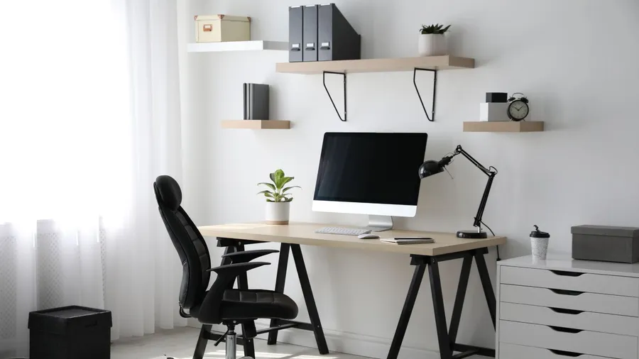 A minimalist home office with a wooden desk with a computer, lamp, and plant, floating shelves, and a chair placed near a window.