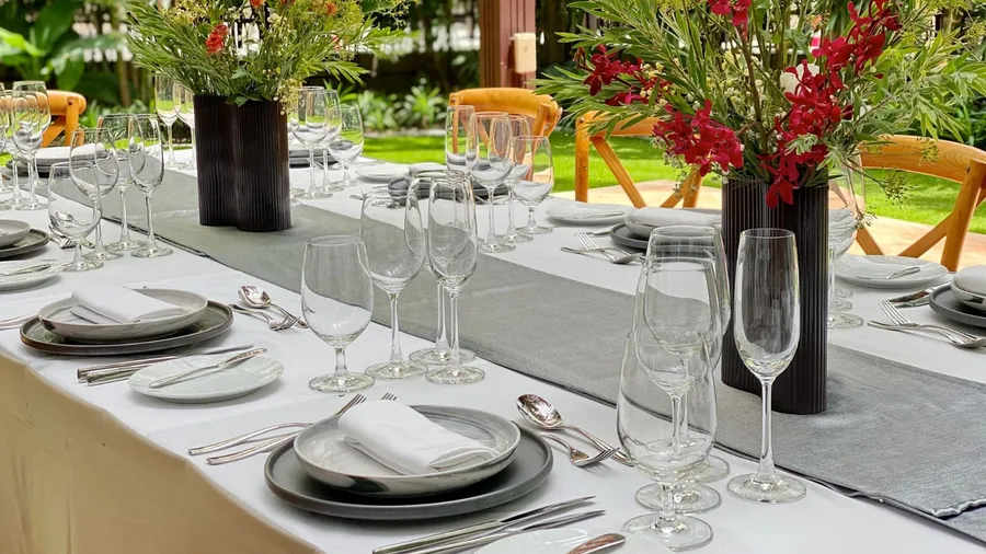 An outdoor dining setup with plates, napkins, cutlery, glassware, and flower vase centerpieces.