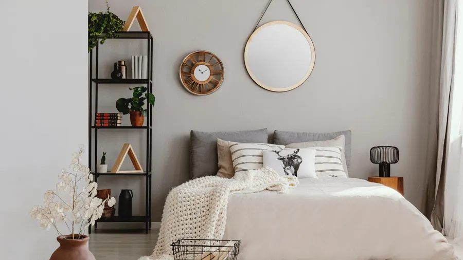 A stylish bedroom interior with a cozy bed with layered pillows and a chunky knit throw , flower vase, shelf with decor pieces, and a round mirror hanging on the wall above the bed.