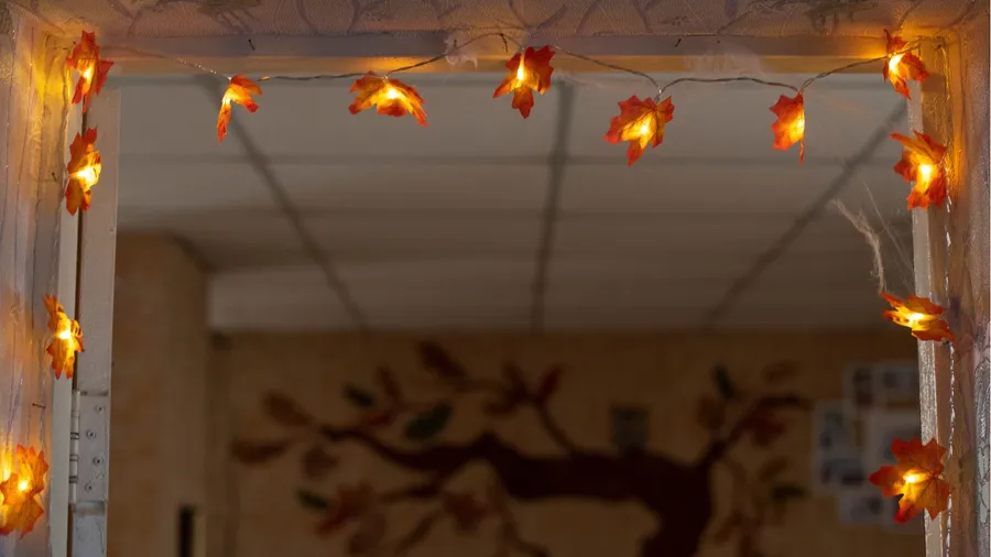 Fall interior decor. Orange electric lights having maple tree leaves shape attached to room door frame. Fall tree painting on the wall seen from door.