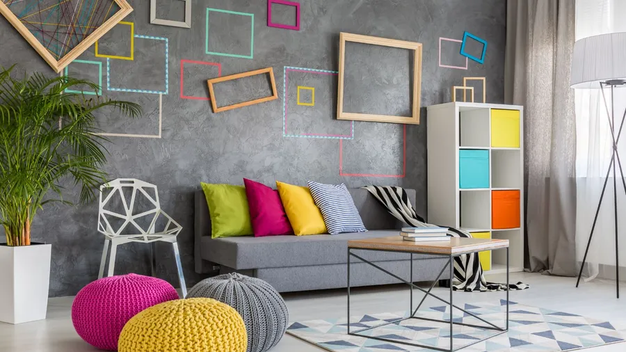 A colorful living room interior with a cozy gray couch with colorful throw pillows, floor cushions, patterned rug, chair, shelf, plant, floor lamp, and colorful rectangular frames on the wall.