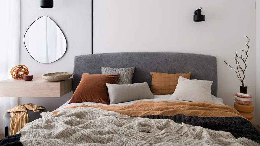 A stylish bedroom interior with a cozy bed, wooden floating nightstand, vase with dried branches, and a unique mirror on the wall.