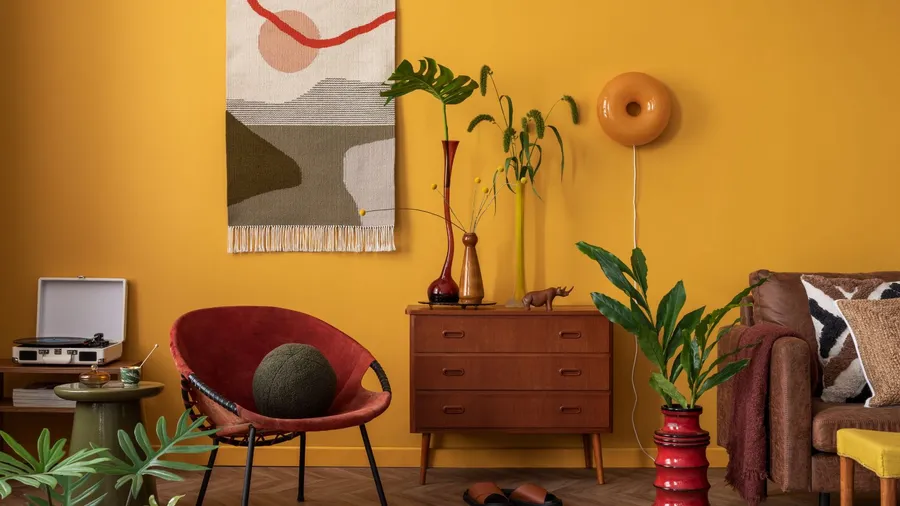 A cozy living room corner with mustard yellow walls, a wooden dresser, a large leafy plant, a brown couch, and decorative items.